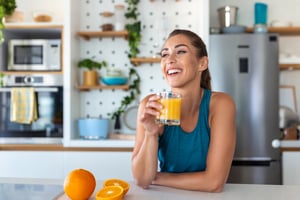 drinking fresh orange juice in kitchen. Healthy diet. Happy young woman with glass of juice and orange at table in kitchen